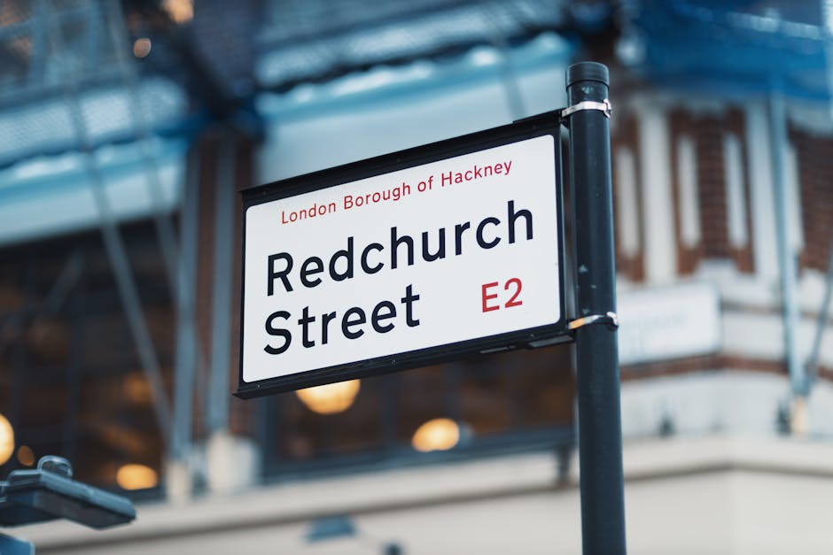 A street sign reading 'Redchurch Street' in the London Borough of Hackney, E2, mounted on a black pole with a white background and black and red lettering, situated in front of a building with large glass windows and brick walls. The area appears well-maintained with clear, clean signage and ambient lighting, indicating a bustling urban commercial or residential district. The focus is on the sign, with a blurred background showing part of a building with a canopy and warm lighting, suggesting a lively, well-kept environment suitable for surface cleaning and maintenance discussions, as highlighted by Carpet Cleaning Hackney on their webpage about insider tips for carpet cleaning near Broadway Market Hackney.