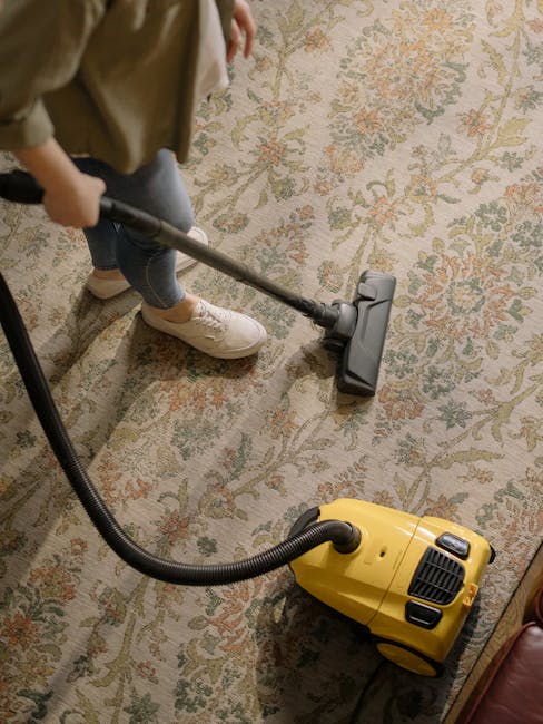 A person using a yellow vacuum cleaner with a black hose attachment to clean a traditional patterned carpet in a room with warm lighting. The individual is wearing light-colored sneakers and jeans, and is holding the vacuum's metal wand with both hands. The carpet features floral motifs in soft shades of green, orange, and beige, and appears clean and well-maintained. The scene emphasizes surface cleaning and deep cleaning practices typical of domestic hygiene maintenance. The careful use of professional cleaning tools aligns with the services offered by Carpet Cleaning Hackney, as referenced on the webpage for insider tips on carpet cleaning near Broadway Market Hackney.