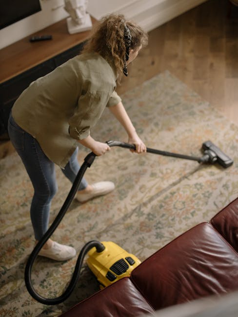 A woman with curly hair, wearing a beige jacket, dark jeans, and white sneakers, is performing surface cleaning with a yellow vacuum cleaner equipped with a hose attachment on a patterned rug in a living room. The room features a wooden floor, a wooden sideboard with decorative items, and a brown leather sofa. The lighting is warm and natural, emphasizing the cleanliness and freshness of the carpet. This image illustrates professional domestic cleaning services offered by Carpet Cleaning Hackney for maintaining hygiene and deep-cleaning carpets in residential spaces, aligning with the 'Quick carpet cleaning guide for Mare Street Hackney, Hackney' page on their website.
