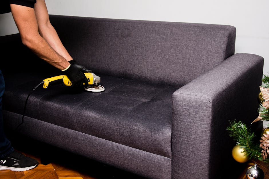 A person using a handheld steam cleaner to deep clean and sanitise a dark grey fabric sofa in a residential living room. The sofa features a modern design with straight lines, wide armrests, and a plush backrest, positioned against a white wall. The cleaning tool is pressed against the sofa's seat cushion, with the operator's hands visible wearing black gloves. The room has wooden flooring, and nearby, a decorated Christmas tree with ornaments and tinsel adds a festive touch. Natural light illuminates the scene, highlighting the sofa's clean and fresh appearance, as part of surface cleaning and hygiene maintenance carried out by Carpet Cleaning Hackney.